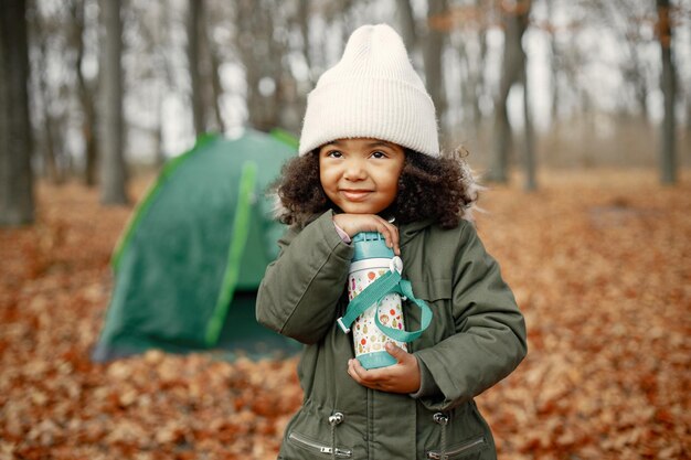 One little black girl in tent camping in the forest Girl is holding a thermos in her hands Black girl wearing khaki coat and beige hat