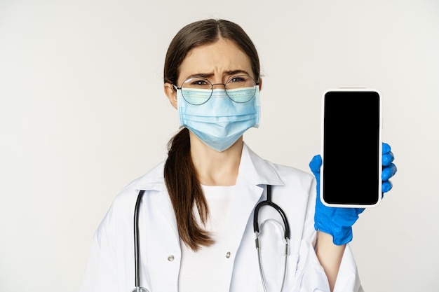 Free photo online medical help concept sad and gloomy young doctor in face mask showing smartphone screen with ...