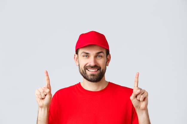 Online shopping, delivery during quarantine and takeaway concept. Cheerful bearded smiling courier in red uniform cap and t-shirt, invite take look at promo, pointing fingers up, grey background