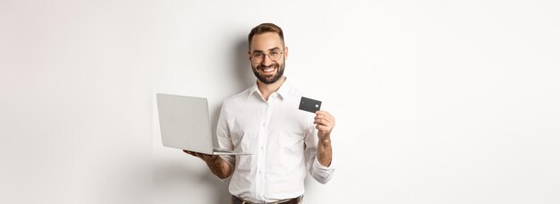 Online shopping handsome man showing credit card and using laptop to order in internet standing over