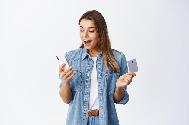 Online shopping. Portrait of excited young woman reading smartphone screen and holding plastic credit card, paying for internet purchase, making an order, white