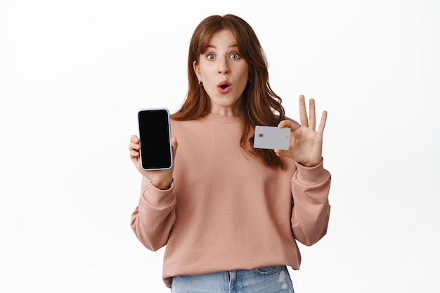 Free photo online shopping. redhead woman gasp amazed, showing smartphone screen and credit card, say wow and look excited, standing over white background