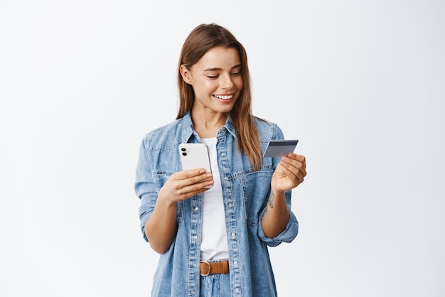 Online shopping Smiling goodlooking woman paying for order using plastic credit card to pay with mobile phone standing against white background