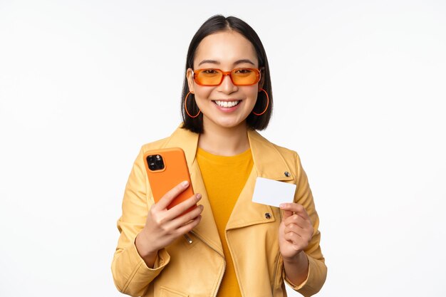 Online shopping Stylish asian female model in sunglasses holding credit card and mobile phone smiling happy standing over white background