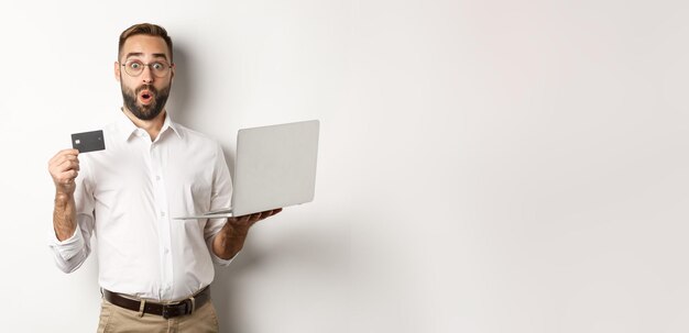 Online shopping surprised man holding laptop and credit card shop internet store standing over white