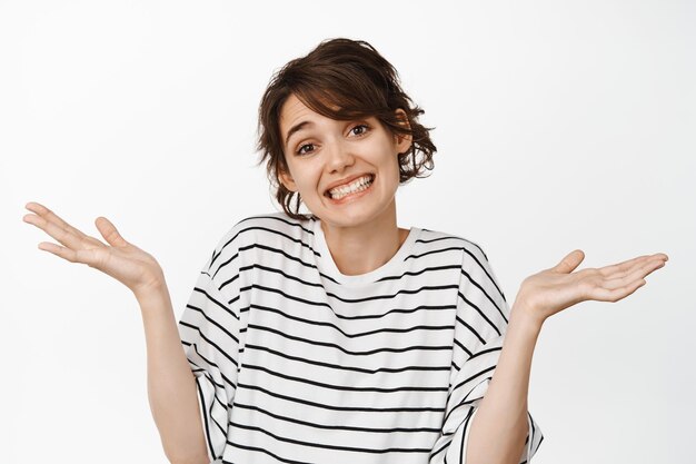 Oops sorry my mistake. Cute awkward woman shrugging shoulders and smiling unaware, cant say, nothing to answer, standing over white background