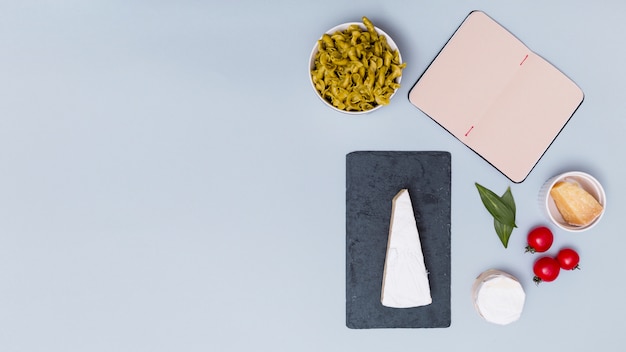 Open blank book and uncooked pasta with ingredient over grey backdrop