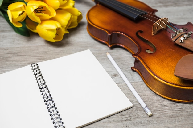 Free photo an open blank notebook; pencil; tulip and classical violin on plank backdrop