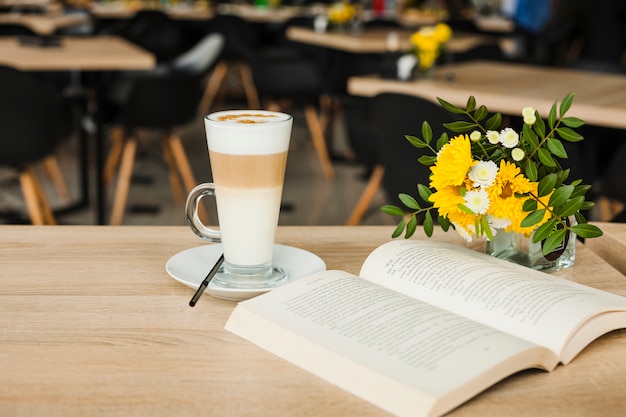 Open book with latte coffee cup and fresh flower vase over wooden table