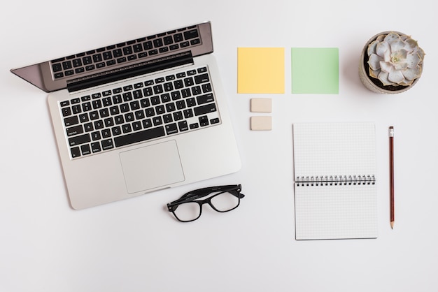 An open laptop; adhesive note; cactus plant; spiral notepad; pencil and eyeglasses on white background