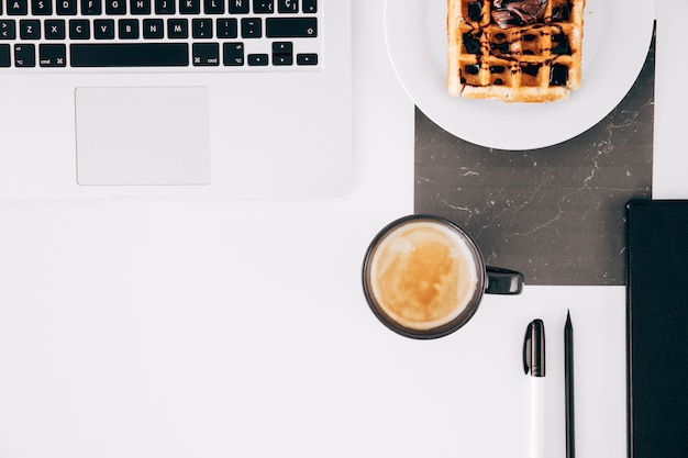 An open laptop; waffle; coffee cup; pencil; pen and open laptop on white desk