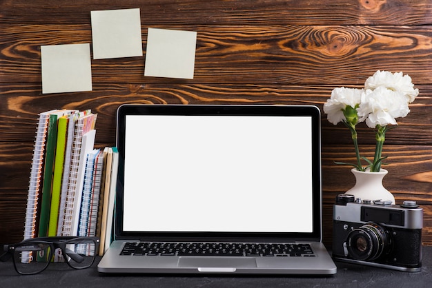 An open laptop with blank white screen; vintage camera; eyeglasses and books on wooden desk