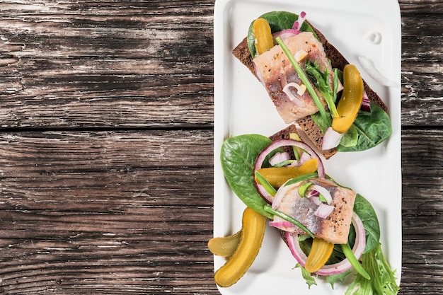 Free Photo open sandwiches with young herring, herbs and pickled cucumber on a white plate, top view with copy space. traditional danish smorrebrod. sandwiches with herring are arranged on an old wooden table