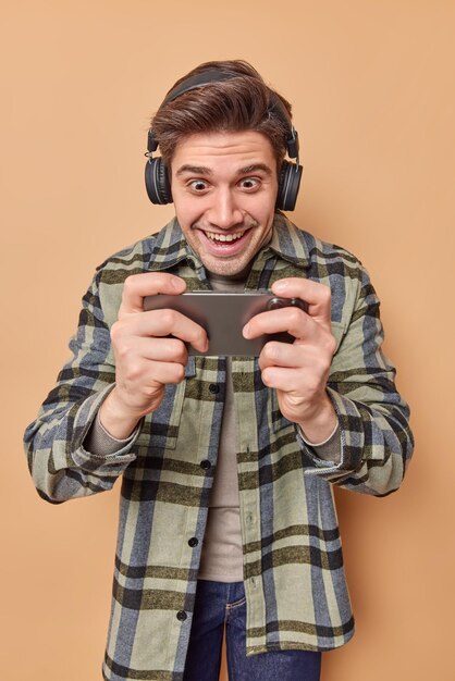 Optimistic addicted man gamer plays video games on smartphone stares at screen of cellular feels glad wears wireless headphones on ears dressed in checkered shirt isolated over beige background