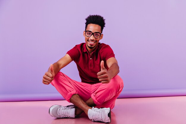 Optimistic african man in pink pants sitting with legs crossed. Smiling smart guy posing on the floor.