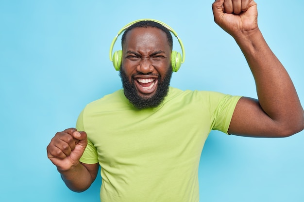 Optimistic carefree bearded man dances with rhythm of music dressed in green t shirt listens music isolated over blue wall