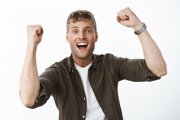 Free photo optimistic and excited charismatic blond guy with blue eyes and white broad smile raising clenched fists in triumph support and cheer smiling delighted from success and victory over gray wall