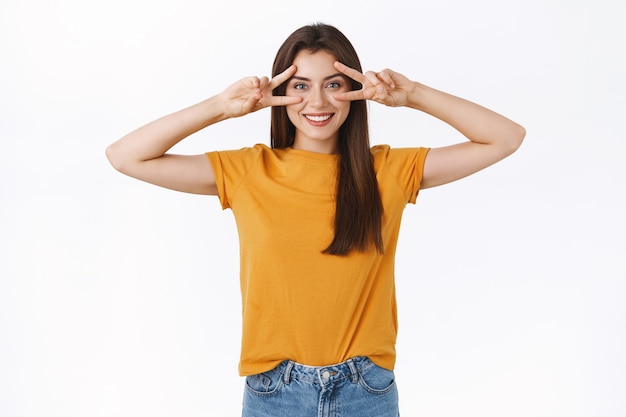 Optimistic, lucky cheerful brunette woman in yellow t-shirt, showing peace or victory sign over eyes, smiling express positivity and joy, enjoy party, like attend amazing event, white background