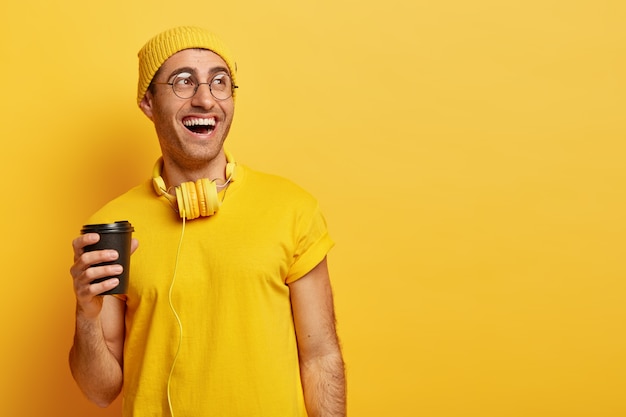 Free Photo optimistic man in glasses laughs as spends time with friends during coffee break, holds disposable cup, looks away