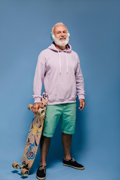 Optimistic man in shorts and hoodie posing with skate board Fashionable guy with beard in blue headphones and modern outfit smiling