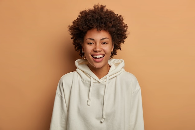 Optimistic teenage girl dressed in casual white sweatshirt, smiles happily, stands against brown space, hears funny joke from friend