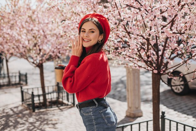 Optimistic woman in bright outfit is smiling cute near sakura. Pretty lady in red sweater anf hat posing in good mood in city garden