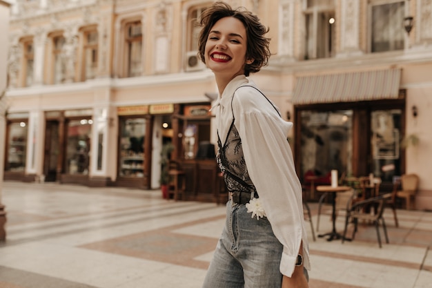 Free photo optimistic woman with curly hair in jeans smiling sincerely in city. cool lady in light blouse with black lace at street.