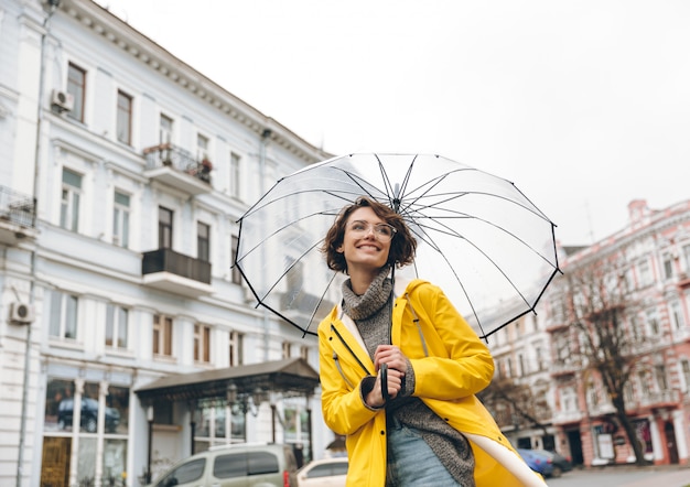 Free Photo optimistic woman in yellow raincoat and glasses having fun while walking through city under big transparent umbrella during cold rainy day