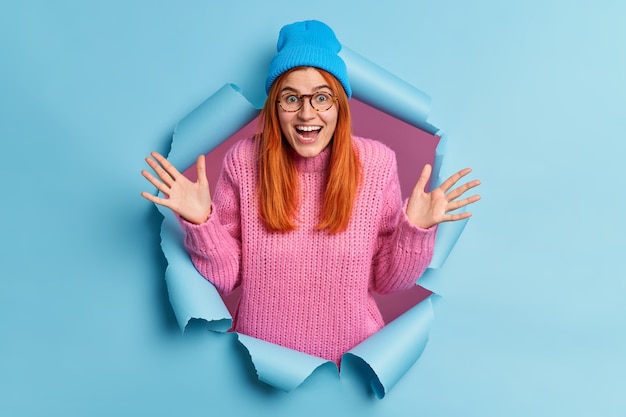 Optimistic young redhead woman feels impressed and fascinated by something raises palms smiles broadly reacts on exciting news has overemotive happy expression.