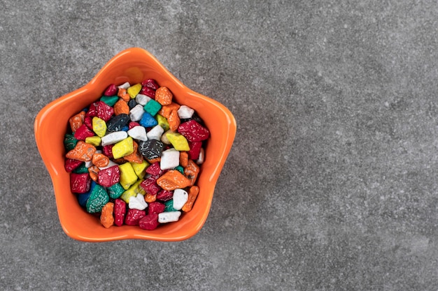 Orange bowl of colorful stone candies on stone table.