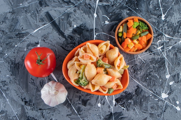 Orange bowl of tasty seashell pasta with vegetable salad on marble surface. 