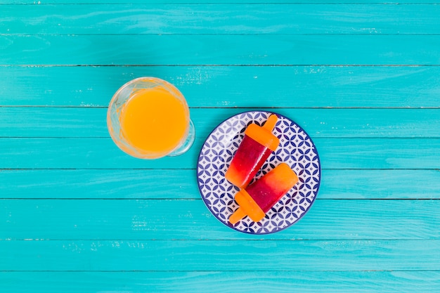 Orange juice and bright fruit popsicle on plate on wooden surface