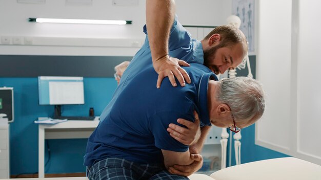 Osteopath helping senior man to crack back bones in cabinet, doing physical therapy exercise. Male assistant using physiotherapy procedure to increase mobility and treat patient.