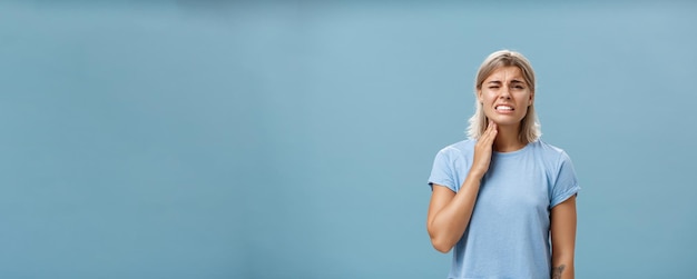 Free photo ouch my throat displeased unhappy goodlooking adult blonde girl with short haircut clenching teeth from painful feeling touching neck having angina or inflammation posing over blue background