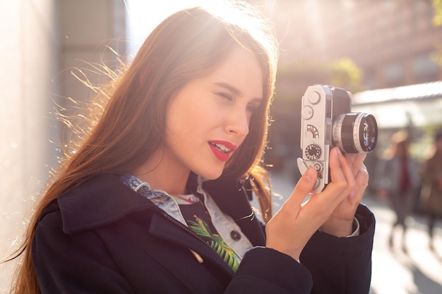 Free photo outdoor autumn smiling lifestyle portrait of pretty young woman, having fun in the city with camera, travel photo of photographer. making pictures in hipster style. sun flare