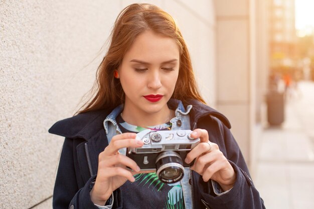 Outdoor autumn smiling lifestyle portrait of pretty young woman, having fun in the city with camera, travel photo of photographer. Making pictures in hipster style. Sun flare