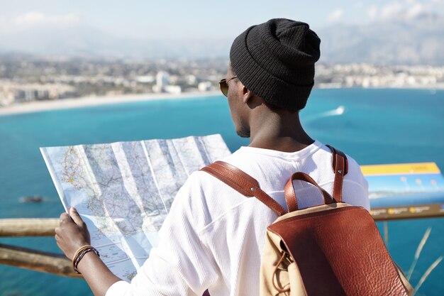 Outdoor back shot of dark-skinned tourist with leather backpack on his shoulders holding paper guide in his hands, facing amazing beautiful view of European sea coast, standing on sightseeing platform