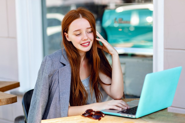 Outdoor city fashion portrait of young businesswoman working at cafe on terrace at sunny day, casual stylish outfit, mint details, using her laptop, cafe break, business concept.