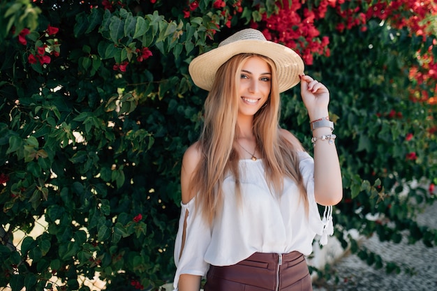 Outdoor close up portrait of young beautiful happy smiling curly girl wearing stylish straw hat in street near blooming roses. Summer fashion concept. Copy space