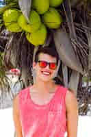 Free photo outdoor fashion portrait of handsome stylish guy spending great time at tropical beach, posing near coconut palm, wearing bright outfit and neon sunglasses.