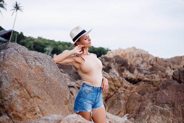 Outdoor fashion portrait of young european woman in beige body bodysuit blue jean shorts and classic stylish hat on tropical rocky beach warm sunset light