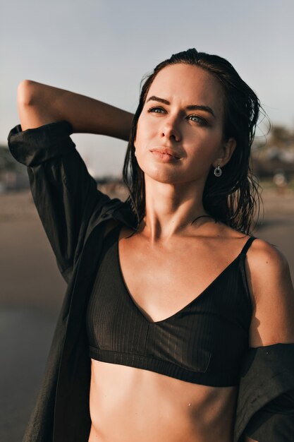 Outdoor fashionable portrait of attractive pretty woman with dark hair and blue eyes looking on the ocean in sunlight