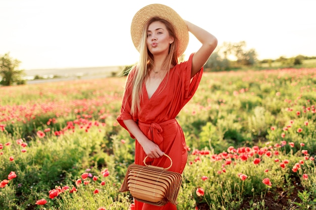 Outdoor fashionable portrait of stunning blonde woman  posing while walk in  amazing poppy field in warm summer evening.  Wearing straw hat, trendy bag and red dress.