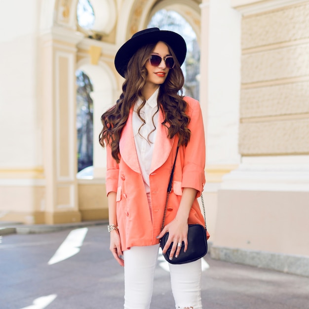 Outdoor hight fashion portrait of stylish casual woman in black hat, pink suit, white blouse posing on old street