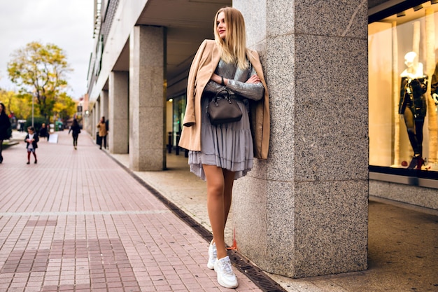 Outdoor lifestyle fashion portrait of pretty elegant glamour blonde woman with long legs, wearing trendy sneakers, dress sweater and coat, posing at Europe city, traveling alone.