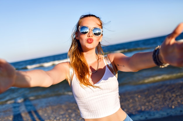 Outdoor lifestyle portrait of funny happy girl traveling to the ocean alone, making selfie at the beach, happy positive emotions, mirrored sunglasses, white crop top and backpack, joy, motion.