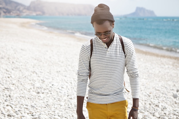 Outdoor lifestyle portrait of stylish young man with backpack and sunglasses walking on sunny European beach, wearing perplexed smile, looking down at pebble