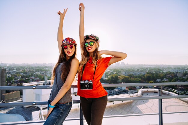 Outdoor lifestyle portrait of two pretty stylish best friends posing on the roof with amazing view to the city, put their hands to the air screaming laughing going crazy and enjoy their freedom