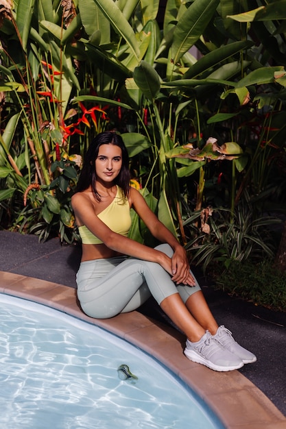 Outdoor portraif of fit slim sporty tanned pretty caucasian woman in sport yellow top and leggings over tropical plants