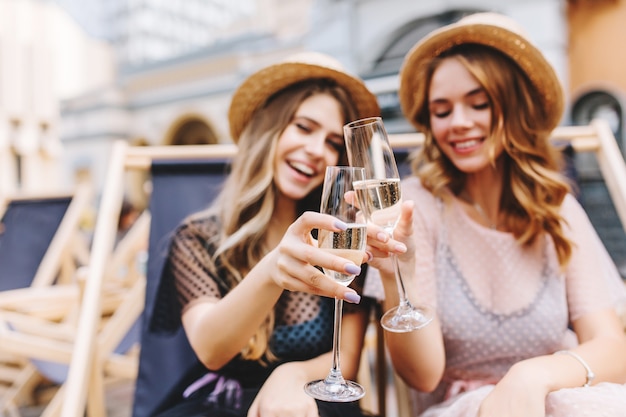 Outdoor portrait of cheerful young women celebrating vacation with goblets of champagne on foreground